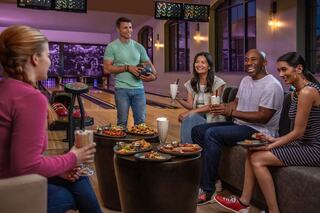 A group of friends enjoys bowling and food, laughing together while holding drinks in a vibrant, cozy bowling alley setting.
