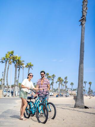 Two friends stand by teal bicycles on a sunny beach, surrounded by palm trees and a clear blue sky, enjoying a leisurely day.