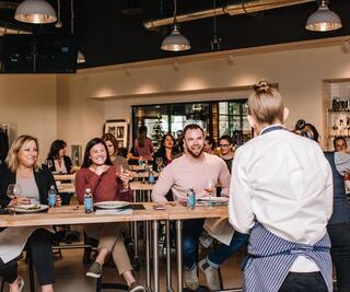 A cheerful group in a modern space enjoys a cooking class, engaging with a chef while savoring food and drinks. Laughter and camaraderie abound.