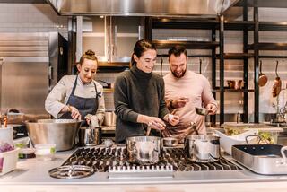 Three people are joyfully cooking together in a modern kitchen, surrounded by pots, pans, and fresh ingredients.