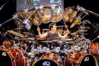 A drummer performs in front of an elaborate drum setup, surrounded by numerous cymbals and instruments, showcasing impressive skill and technique.