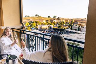 Two women in plush robes toast with champagne on a balcony overlooking a lively outdoor event and scenic surroundings.