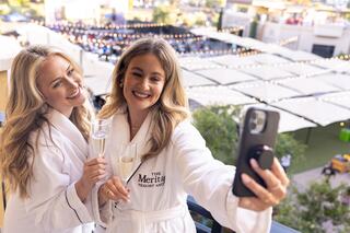 Two women in bathrobes take a cheerful selfie on a balcony, holding glasses of champagne, with festive lights in the background.