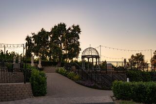 A serene garden setting at dusk features a gazebo, lush greenery, and decorative string lights, creating a tranquil atmosphere.