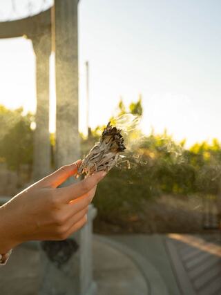 A hand holds burning sage, releasing fragrant smoke against a warm sunset backdrop with greenery. Tranquil and calming scene.