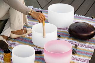 A person uses a mallet on a crystal singing bowl, surrounded by various instruments on a colorful woven blanket. Soft, serene atmosphere.