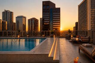 A rooftop pool glows as the sun sets behind modern skyscrapers, creating a peaceful urban oasis with cozy seating and drinks.