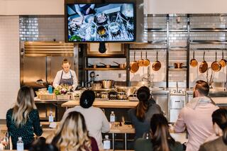 A cooking demonstration is underway, with a chef presenting in a modern kitchen while an audience observes attentively.
