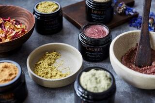 A collection of vibrant natural powders and dried petals, alongside jars of skincare products, arranged on a textured surface.