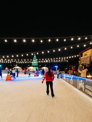 A skater in a red sweater glides on ice at night, surrounded by twinkling lights, a festive tree, and people enjoying the rink.