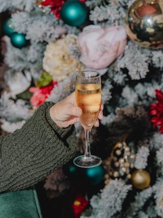 A hand holds a champagne flute filled with bubbly, set against a festive backdrop of decorated Christmas tree branches and ornaments.