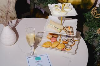 A tiered stand displays an assortment of pastries and sandwiches, accompanied by a glass of champagne and a menu on a stylish table.