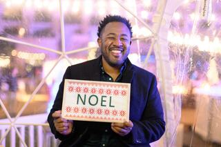 A smiling man holds a festive sign that reads "NOEL," surrounded by bright lights in a cozy, decorated space.