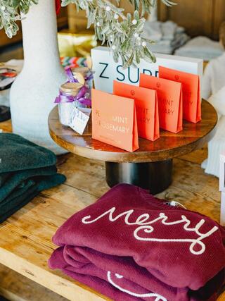 Cozy holiday items are displayed on a wooden table, featuring sweaters, greeting cards, and a decorative jar amidst festive decor.
