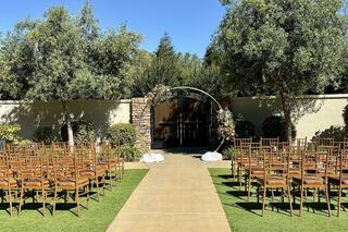 A beautifully arranged outdoor wedding ceremony with rows of chairs, a decorative arch, and lush greenery under a clear blue sky.