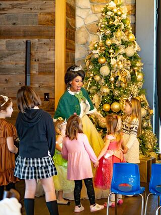 A character in a festive green gown engages with a group of children, surrounded by a beautifully decorated Christmas tree.