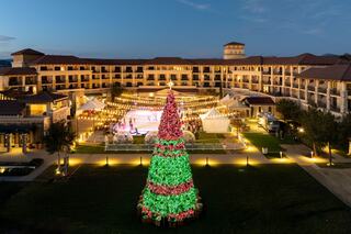 A beautifully lit holiday tree stands in front of a hotel, with an ice skating rink and festive decor illuminating the evening sky.