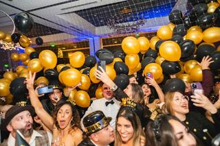 A lively celebration filled with people wearing festive hats, surrounded by black and gold balloons, capturing the joyous New Year's spirit.