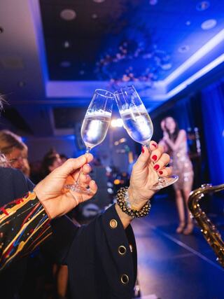 Two elegantly dressed hands toast with champagne flutes, celebrating against a festive backdrop with a performer in the distance.