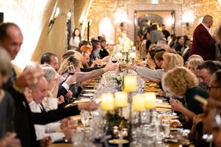 A long communal dining table filled with people celebrating, raising glasses in a toast, adorned with candles and flowers.