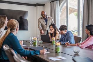 A diverse group collaborates around a conference table, engaged in discussion as a leader presents insights using a laptop.
