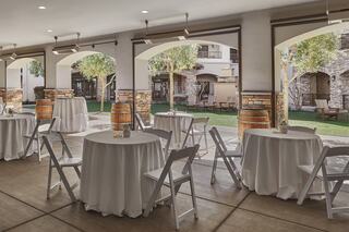 An elegant outdoor setting features round tables with white tablecloths, wooden barrels, and a view of a green courtyard with trees.