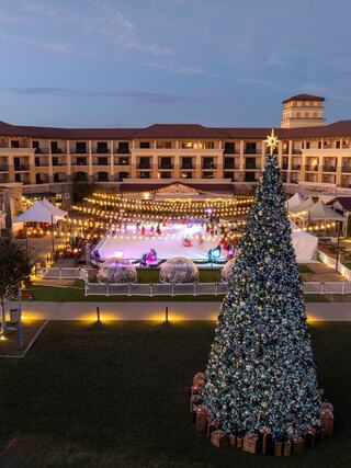 A festive scene features a large decorated Christmas tree near an outdoor ice rink, illuminated with string lights, surrounded by cozy tents.