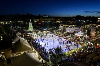 A bustling outdoor ice skating rink is surrounded by festive lights, a Christmas tree, and crowds enjoying the evening atmosphere.