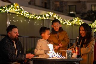 A family enjoys a cozy evening by the fire, roasting marshmallows, with festive lights twinkling in the background.