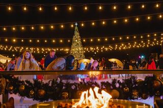 A festive scene features a sparkling Christmas tree amid bright lights, with people enjoying the atmosphere and a cozy fire in the foreground.