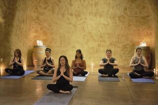 A group of six women practice yoga in a serene setting, seated cross-legged on mats with hands in a prayer position, surrounded by soft candlelight.