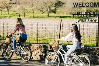 Two women ride bicycles through a vineyard, enjoying a sunny day. A welcoming sign for wine-related experiences is visible nearby.