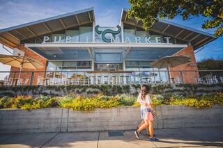 A woman strolls past a modern public market building adorned with large glass windows and vibrant landscaping under a sunny sky.