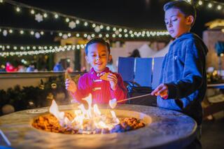 Two children enjoy roasting marshmallows over a fire pit, surrounded by twinkling lights and a festive atmosphere on a cool evening.