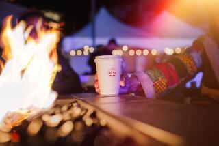 A cozy scene with a hand holding a warm drink near a crackling fire, surrounded by soft lighting and festive ambiance.
