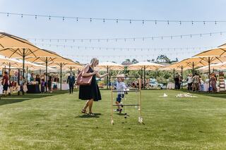 A woman plays with a child on a grassy area, surrounded by market stalls and bright umbrellas under a clear blue sky.