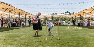 A cheerful scene at an outdoor market with umbrellas, where a woman encourages a young child as they play on lush green grass.