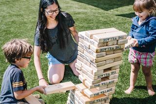 A woman and two children joyfully play a game, stacking wooden blocks in a grassy area, focused on not toppling the tower.