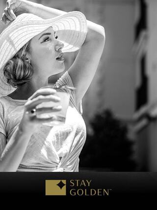 A stylish woman in a sun hat enjoys a drink, exuding elegance and a carefree vibe in a sunny outdoor setting.
