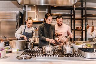 Three people joyfully cook together in a modern kitchen, smiling and engaging with various pots and ingredients on the counter.