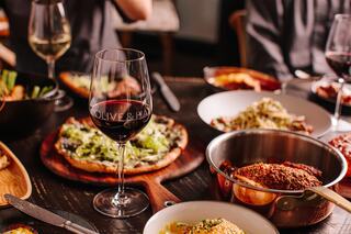 A table spread features a glass of red wine, pizza topped with veggies, and various delicious dishes, creating a cozy dining atmosphere.