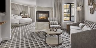 A stylish hotel suite features a cozy seating area, modern furnishings, a fireplace, and large windows with elegant drapes.