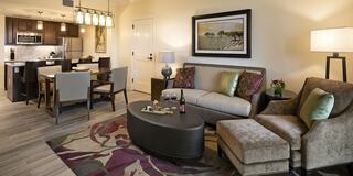 A cozy, modern hotel suite living space featuring a comfortable sofa, dining area, stylish decor, and a well-equipped kitchen. Warm lighting enhances the ambiance.