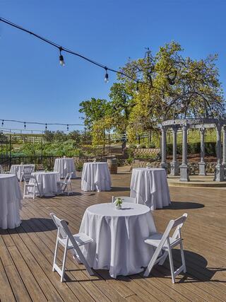 A sunlit outdoor vineyard venue features round tables with white tablecloths and wooden flooring, adorned with a decorative gazebo and string lights.