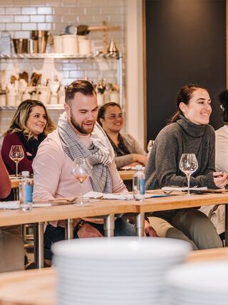 A group of people enjoying a culinary class, sharing laughter and conversation while seated at a communal table with wine glasses.