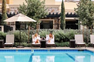 Two girl friends sitting in pool chairs by the pool wearing white robes and drinking wine