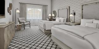 A stylish luxury hotel room features a king-sized bed, elegant decor, a work area, and large windows, creating a bright and inviting atmosphere.