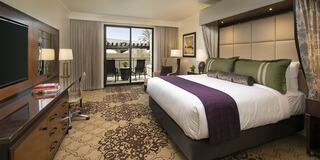 A cozy luxury hotel room features a large bed with plush pillows, a desk, lamps, and a balcony overlooking the scenery. Elegant decor enhances the ambiance.