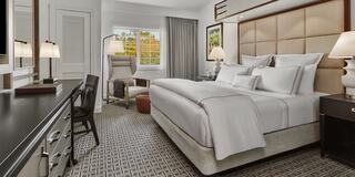 A stylish luxury hotel room features a king-sized bed, elegant decor, a work area, and large windows, creating a bright and inviting atmosphere.