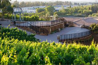 A scenic vineyard features lush green grapevines, elegant pathways, and a charming gazebo overlooking a beautifully landscaped area.